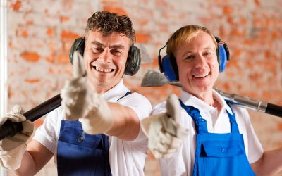 Two construction workers standing in a construction site in front of a brick wall. Their are wearing ear protection and having the thumps up